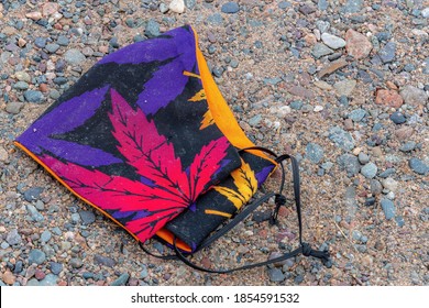 A mask with red and purple cannabis leaves. Mask is lying on gravel. This is the type of mask people use to help prevent the spread of coronavirus.