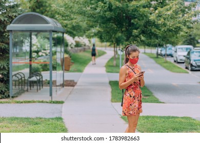 Mask Wearing Woman Waiting For The Bus Holding Mobile Phone COVID-19 Contact Tracing App When Using Public Transport. Obligatory Face Covering Wear In Many Cities As Corona Virus Prevention.