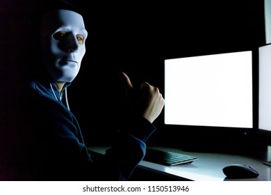 Masked And Anonymous Hacker Under Hood Giving Thumbs Up While Using A Computer With Empty Blank White Screen In The Dark Room - With Copy Space On Screen