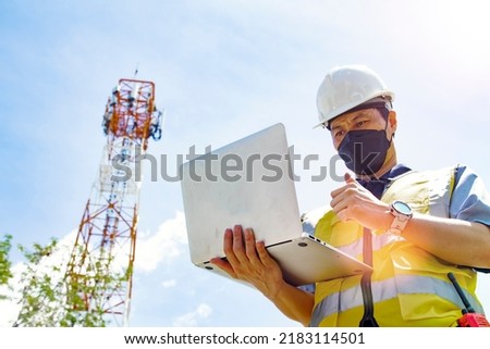 Masked asian male electrical engineer architect uses laptop to field work near telecommunication tower to inspect 5Gg telecommunication technology network tower network thumbs up confidently.