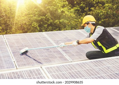 Masked Asian Male Worker Wearing A Mask And Helmet Is Cleaning A Blue Outdoor Solar Panel Using A Dirt Brush For Effective Use : Cleaning With A Brush And Inspecting Solar Panels On Industrial Roofs