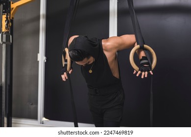 A masked asian man looking down while leaning on wooden still rings. A gymnast preparing to train at the gym. - Shutterstock ID 2247469819