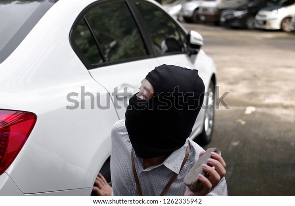 Masked\
burglar wearing a balaclava holding little stone ready to burglary\
against car background. Insurance crime\
concept.