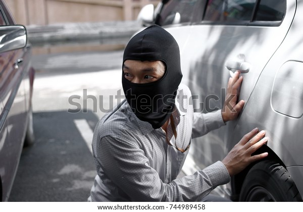 Masked burglar\
wearing a balaclava ready to burglary against car background.\
Insurance crime concept.