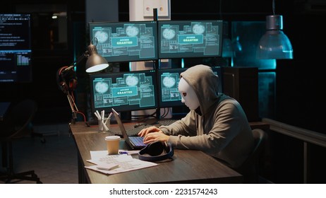 Masked criminal hacking computer server at night, working on laptop and multiple monitors to steal IT information. Hacker with hidden identity acting dangerous, cloud computing. Handheld shot.