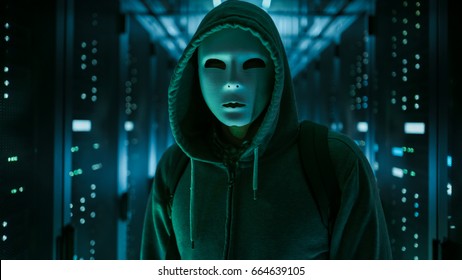 Masked Hacker in a Hoodie Standing in Corporate Data Center with Rows of Working Rack Servers.