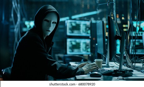 Masked Hacker is Using Computer for Organizing Massive Data Breach Attack on Corporate Servers. They're in Underground Secret Location Surrounded by Displays and Cables. - Shutterstock ID 680078920