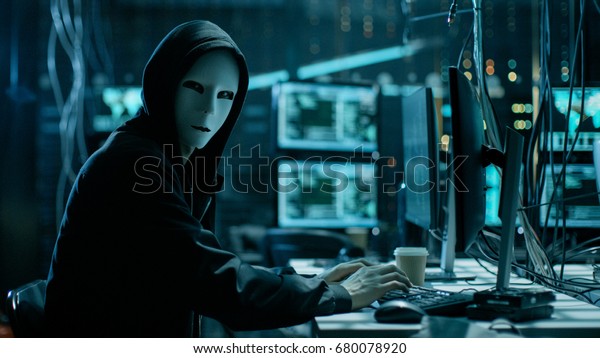 Masked Hacker is Using\
Computer for Organizing Massive Data Breach Attack on Corporate\
Servers. They\'re in Underground Secret Location Surrounded by\
Displays and Cables.