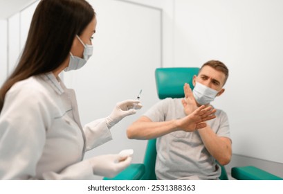 Masked healthcare professional prepares to administer vaccine, while nervous male patient raises his hands in apprehension. The image captures vaccine anxiety and healthcare in a clinical setting - Powered by Shutterstock