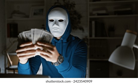 Masked man in hoodie holding us dollars at night in a dimly lit office, suggesting illicit financial activity.