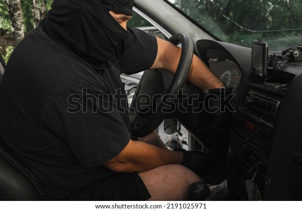 A masked robber steals a\
car by starting the car with wires with his hands in black leather\
gloves.