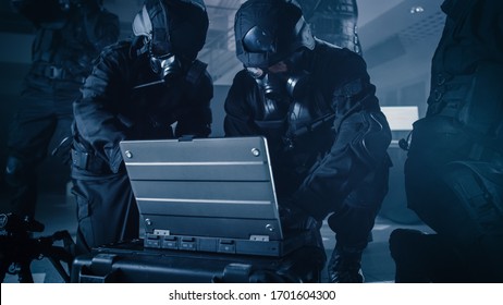 Masked Team Of Armed SWAT Police Officers With Rifles Are In Dark Seized Office Building With Desks And Computers. Soldier Opens A Laptop Computer To Plan A Tactical Attack.