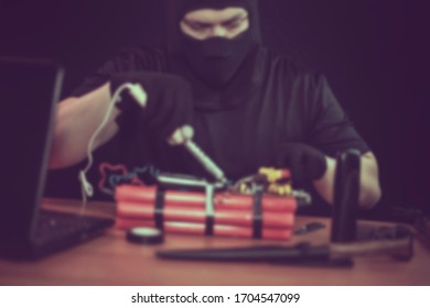 Masked terrorist preparing an explosive device with a soldering iron in hand, indoors on the table: a detonator from a mobile phone, a gun, a knife, a laptop; toned, image is blurry
