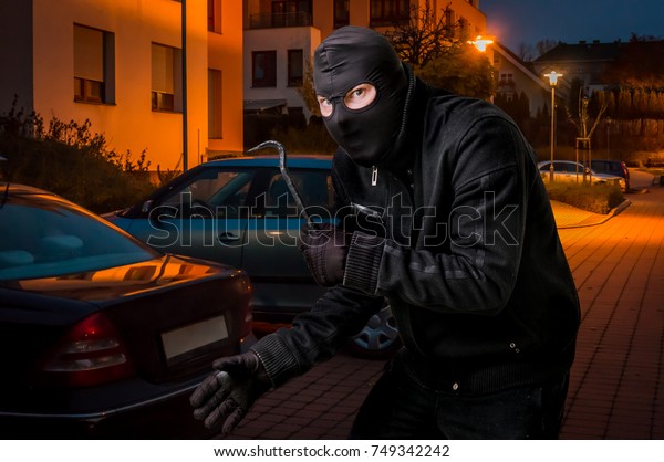 Masked thief in balaclava with crowbar wants to rob\
a car at night