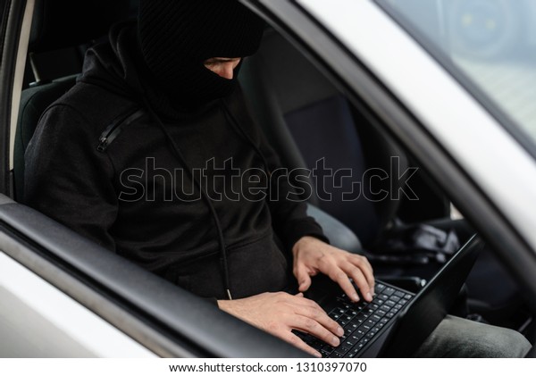 Masked thief hacker in a balaclava\
disarming car security systems and stealing a\
car