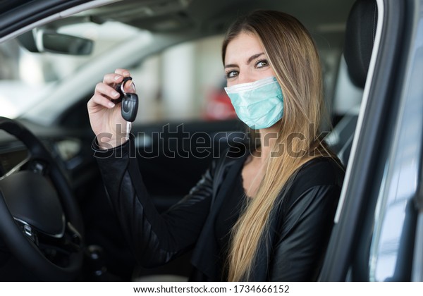 Masked woman showing the key of her\
new car in a car dealer saloon during coronavirus\
pandemic