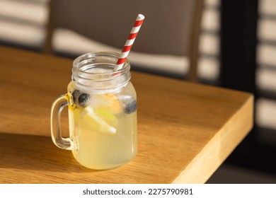 Mason jar glass of fresh lemonade with sliced lemons, lime, orange and blueberries, striped paper straw on cafe table. - Shutterstock ID 2275790981