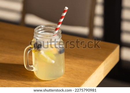 Mason jar glass of fresh lemonade with sliced lemons, lime, orange and blueberries, striped paper straw on cafe table.
