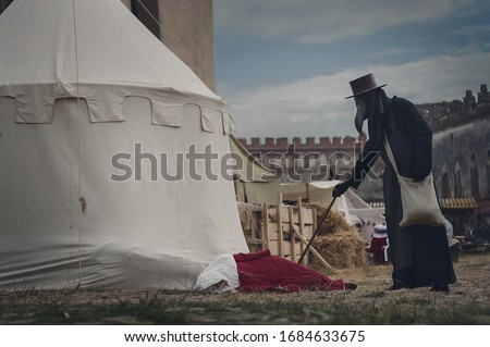 A masquerade historical scene reconstruction. Plague doctor in medieval old town touch a fallen woman. Castle and epidemic of black death