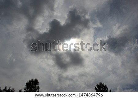 mass of dark gray clouds with sun shining from behind with rays flowing out from behind and tree tops along bottom 