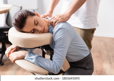Massage therapist massaging shoulders of woman with closed eyes, sitting on massage chair with office on blurred background