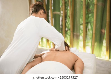 Massage therapist in white outfit does a back massage to a lying woman in cozy salon, back view, selective focus. Body care, treatment, recovery, relax, lymphatic drainage, rebalancing, stress relief. - Powered by Shutterstock