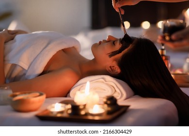 Masseurs Hand Do Holding Glass And Brushing A Facial Mask Herb Black Cream To On Customer's Face As Sleep And Covered Warp By A White On Her Body.