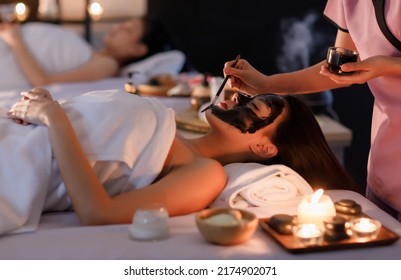 Masseurs Hand Do Holding Glass And Brushing A Facial Mask Herb Black Cream To On Customer's Face As Sleep And Covered Warp By A White On Her Body.