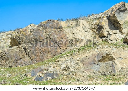 A massive, angular rock formation on a hillside, exhibiting shades of brown, red, and yellow with lichen growth under a bright blue sky.