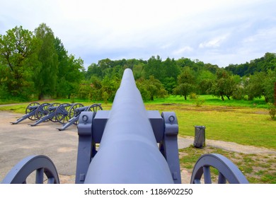 A massive cannon seen from the loading side with two wheels on the sides and three more cannons visible in the distance located on a path near a lawn and a set of trees seen during autumn day