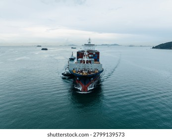 A massive container ship moves through coastal waters under overcast skies assisted by two tugboats in chonburi province thailand.