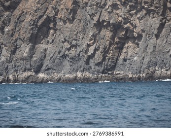 Massive dark cliffside texture dropping into wavy ocean water in bright daylight