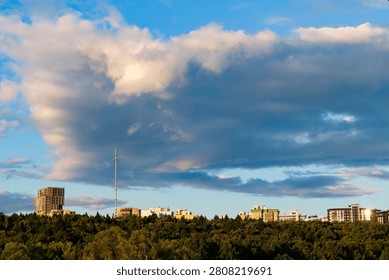 Massive fluffy clouds drift over the modern skyline and a dense green forest, catching the warm golden hour light in a peaceful urban landscape scene