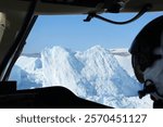 A massive iceberg is seen through the cockpit window of a helicopter flying near the Eqip Sermia Glacier in Greenland, with the pilot’s helmet visible in the foreground