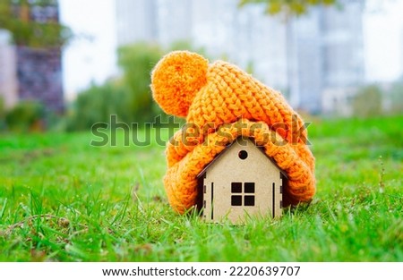 Massive knitted hat on top of a small wooden home model placed on a green lawn with city background. Staying warm before switching central heating on.