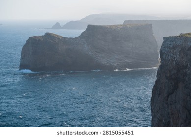 Massive layered cliffs jut into hazy blue Atlantic waters along Portugal's dramatic Algarve coast near Sagres. Waves crash against the towering rock formations beneath a soft, misty morning sky