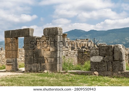 Massive monolithic stone doorframes and lintels stand in the area near the Capitoline Temple at Volubilis, with the Arch of Caracalla and distant mountains visible in the background.