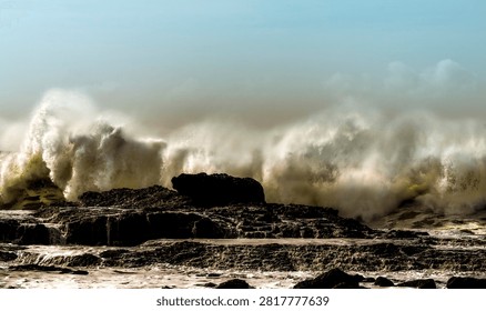Massive ocean waves create dramatic white spray as they break over jagged, dark rocks along the coast