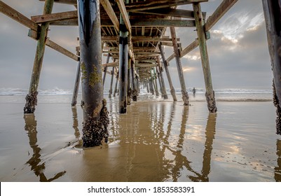 A massive pier overhead frames a lone surfer heading out to the waves near sunset, serving as a kind of seaside sanctuary for those that daily brave the pounding surf. Horizontal. Outdoors. Nature.