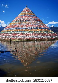 A massive, pyramidal structure completely covered in colorful layered Tibetan prayer flags sitting directly in a calm body of water