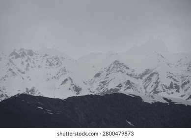 A massive, rugged mountain range dominates the upper portion of the frame, its steep slopes and jagged peaks heavily covered in thick white snow. The towering, high-altitude summits disappear into a dense layer of low-hanging, overcast clouds that completely shroud the upper sky in a muted grey fog.

In stark contrast to the white peaks above, a dark, nearly black mountain ridge stretches horizontally across the foreground, cutting a sharp, shadowed profile against the snowy landscape behind it.
