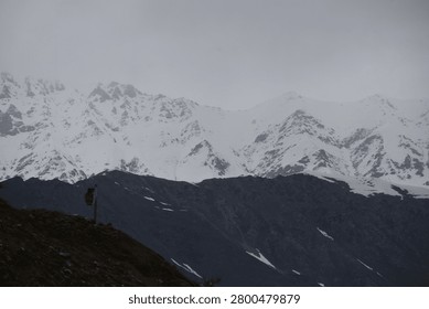 A massive, rugged mountain range dominates the upper portion of the frame, its steep slopes and jagged peaks heavily covered in thick white snow. The towering, high-altitude summits disappear into a dense layer of low-hanging, overcast clouds that completely shroud the upper sky in a muted grey fog.

In stark contrast to the white peaks above, a dark, nearly black mountain ridge stretches horizontally across the foreground, cutting a sharp, shadowed profile against the snowy landscape behind it.