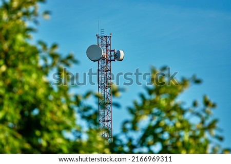 Mast with cell phone antennas against blue sky. Tree with green leaves.
