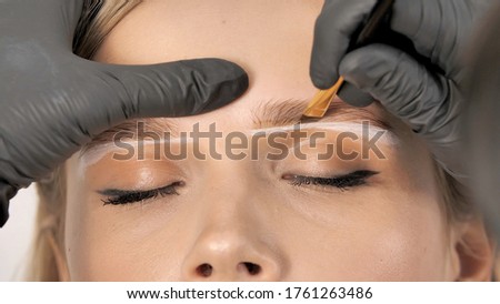 Master applies brow paste with a brush to eyebrows. Beautiful attractive female face of a blonde well-groomed woman or lady. Styling and lamination of eyebrows. Stylist's hands in black gloves