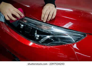 The master applies vinyl film to the headlight of a red car. Closeup view on worker detailer hand smoothing with a scraper protective film.  - Powered by Shutterstock