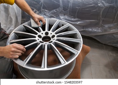 Master body repair man is working on preparing the surface of the aluminum wheel of the car for subsequent painting in the workshop, cleaning and leveling the disk with the help of abrasive material