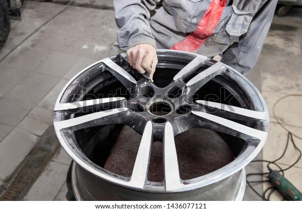 Master body repair man is working on preparing\
the surface of the aluminum wheel of the car for subsequent\
painting in the workshop, cleaning and leveling the disk with the\
help of abrasive\
material