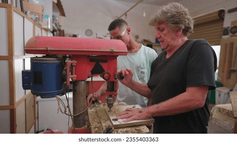 Master carpenter using industrial machine to make a hole into wooden surface while teaching apprentice to use equipment, portrait of Brazilian workers