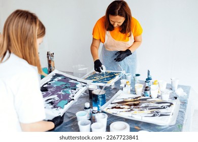 Master class on an Acrylic Fluid Pouring. Young women paint with liquid acrylic in an art workshop. Fluid Art. Dirty glass technique. There are paintings, paints and brushes on the table. Art therapy.