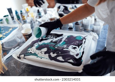 Master class on an Acrylic Fluid Pouring. Teacher and teen girl paint with liquid acrylic in an art workshop. Fluid Art. Dirty glass technique. There are paintings, paints and brushes on the table Art